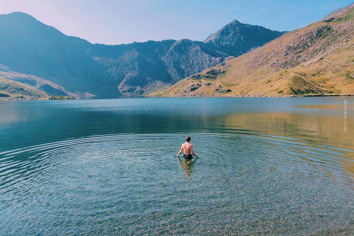 A man stands in the blue water of the lake and the mountains can be seen in the background.