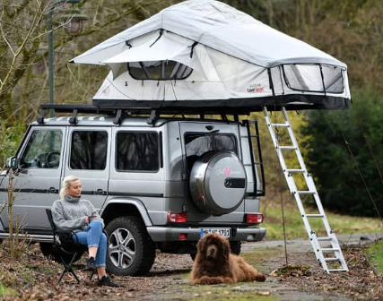 Frau sitzt auf einem Campingstuhl mit ihrem Hund vor ihrem Auto mit Adventure Dachzelt