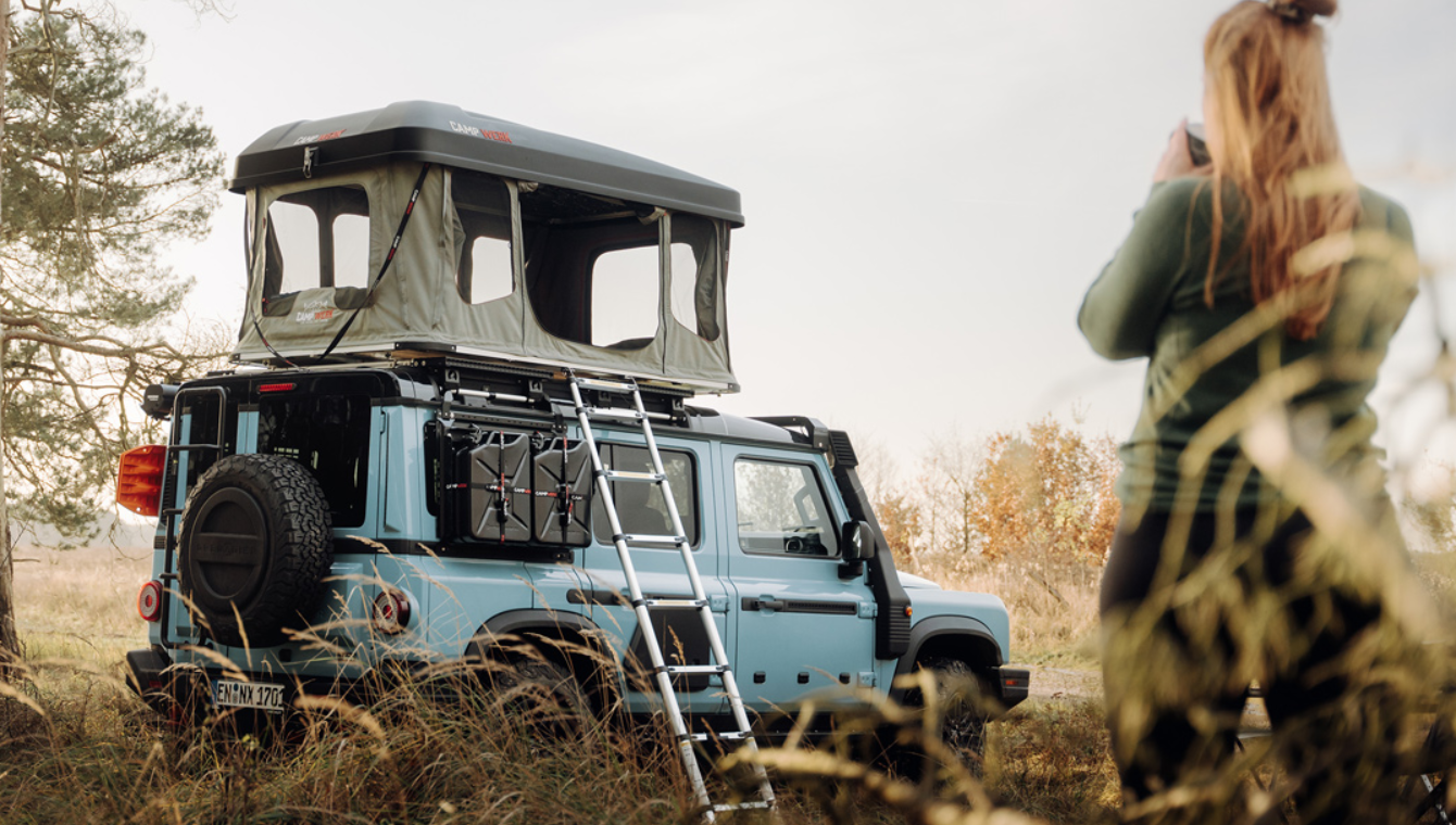 Frau trinkt vor ihrem Auto mit Good Vibes Dachzelt eine Tasse Kaffee