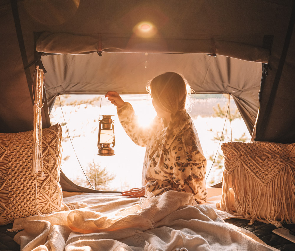 Young woman sits with a camping lamp in the roof tent at sunset
